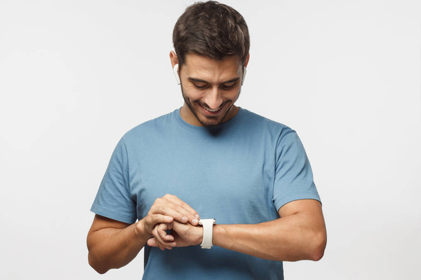 Young handsome man wearing wireless headphones and blue t shirt, checking smart watches with touch screen - Фото, изображение