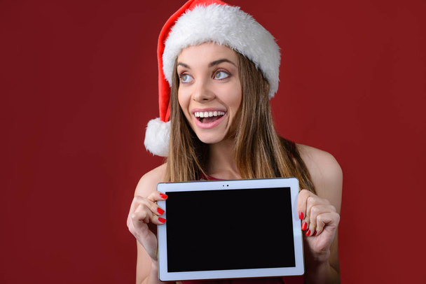 Concept of buying presents on Christmas holidays. Happy impressed woman in red dress and santa hat is holding digital tablet with empty clean black screen for writing text. - Фото, зображення