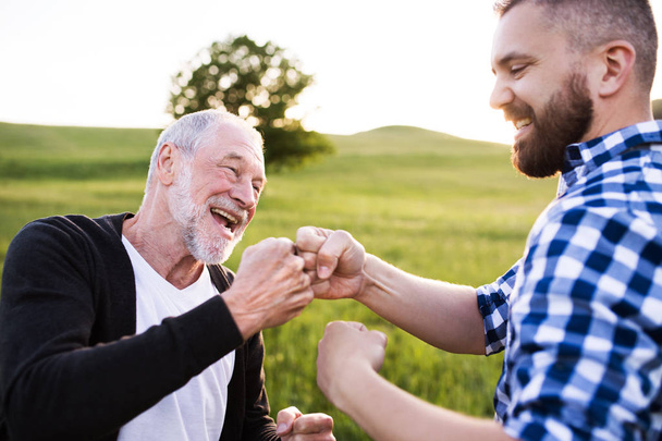 An adult hipster son with senior father making fist bump in nature at sunset. - Φωτογραφία, εικόνα