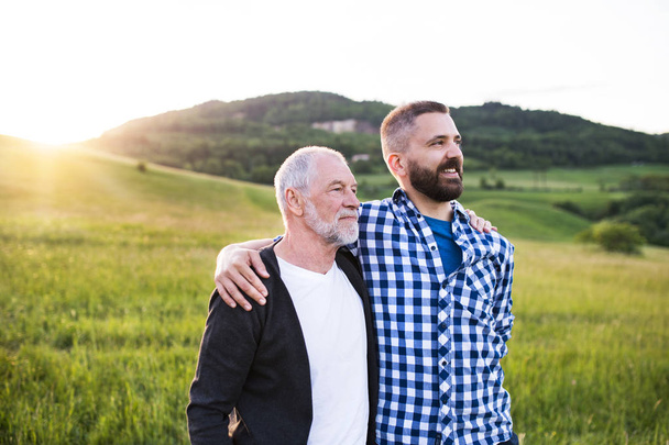 A portrait of an adult hipster son with senior father in nature at sunset, arms around each other. - Foto, Imagen