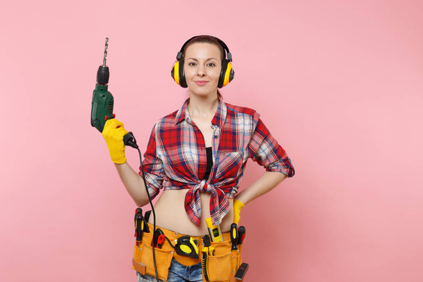 Energy sexy handyman woman in gloves, noise insulated headphones, kit tools belt full of instruments holding power electric drill isolated on pink background. Female in male work. Renovation concept - Photo, Image