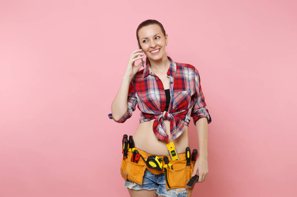 Jonge klusjesman-vrouw in plaid shirt, riem kit tools vol instrumenten praten op mobiele telefoon, uitvoeren van aangenaam gesprek geïsoleerd op roze achtergrond. Vrouw in mannelijke werk. Renovatie concept - Foto, afbeelding