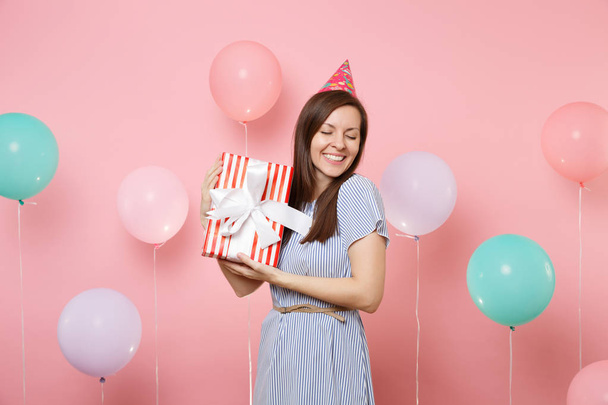 Retrato de mujer alegre con los ojos cerrados en el sombrero de cumpleaños vestido azul que sostiene la caja roja con regalo presente en el fondo rosa con globos de aire de colores. Fiesta de cumpleaños, gente emociones sinceras
 - Foto, Imagen
