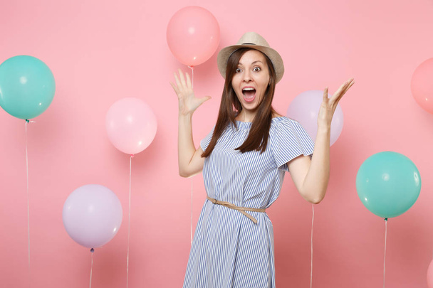 Portrait d'une jeune femme heureuse surprise portant un chapeau d'été en paille et une robe bleue écartant les mains sur un fond rose pastel avec des ballons à air coloré. Anniversaire fête gens émotions sincères
 - Photo, image