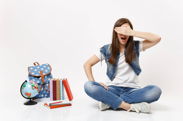 Portrait of young casual woman student screaming covering face with hand and sitting near globe backpack school books isolated on white background. Education in high school university college concept - Photo, Image