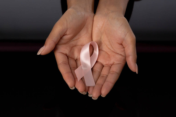 Two hands showing pink ribbon against breast cancer against black background - 写真・画像
