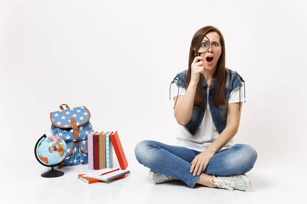 Young shocked irritated woman student holding looking on magnifying glass sitting near globe, backpack, school books isolated on white background. Education in high school university college concept - Photo, Image