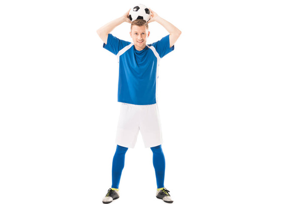 full length view of happy young soccer player holding ball above head and smiling at camera isolated on white - Foto, imagen