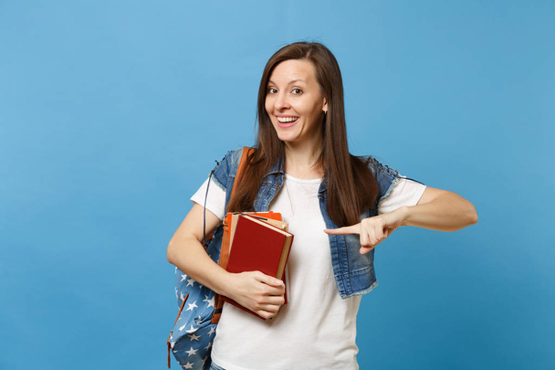 Retrato de una joven estudiante sonriente emocionada con una mochila sosteniendo libros escolares apuntando con el dedo índice al espacio de copia aislado sobre fondo azul. Educación en la escuela secundaria concepto universitario
 - Foto, Imagen