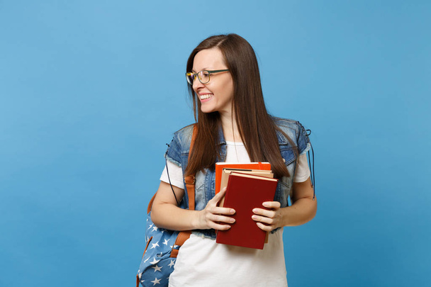 Retrato de joven sonriente atractiva estudiante feliz en gafas con mochila mirando a un lado, sosteniendo libros escolares aislados sobre fondo azul. Educación en la escuela secundaria concepto universitario
 - Foto, imagen