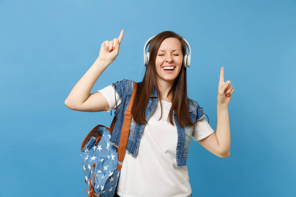Young cheerful woman student with closed eyes with backpack, headphones listen music pointing index finger up dancing isolated on blue background. Education in college. Copy space for advertisement - Foto, Imagen