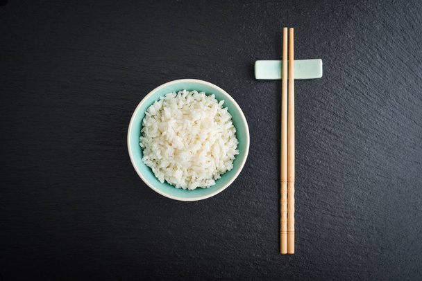 Boiled rice in bowl and  bamboo chopsticks on black slate background, top view - Foto, Bild