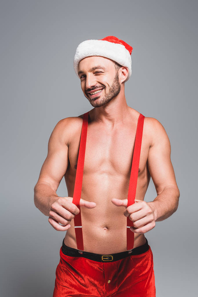 smiling shirtless muscular man in christmas hat winking holding suspenders isolated on grey background - Photo, Image