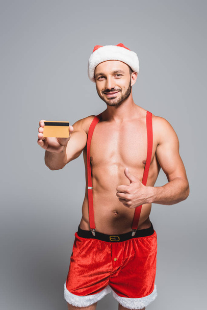 muskularny mężczyzna w Boże Narodzenie kapelusz robi kciuk i wyświetlone karty kredytowej na białym tle na szarym tle - Zdjęcie, obraz