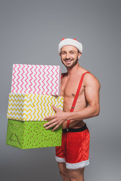 happy muscular man in christmas hat holding pile of gift boxes isolated on grey background - Photo, image