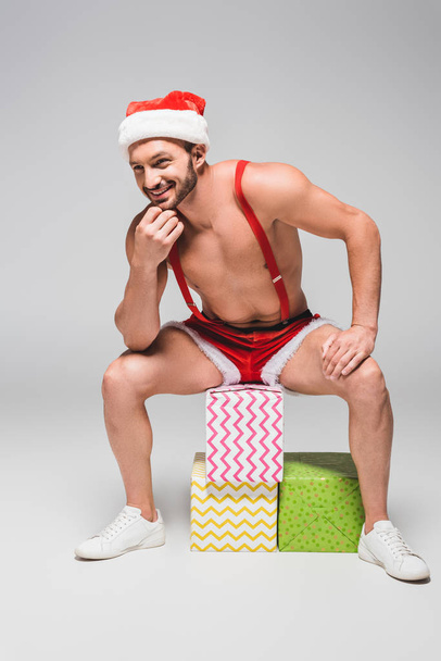 uśmiechający się muskularny mężczyzna w Boże Narodzenie kapelusz i szortach siedzi na pudełka na szarym tle - Zdjęcie, obraz