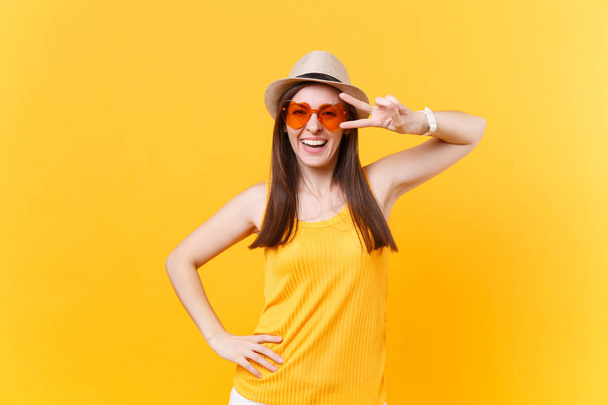 Portrait of smiling excited young woman in straw summer hat, orange glasses showing victory sign, copy space isolated on yellow background. People sincere emotions lifestyle concept. Advertising area - 写真・画像