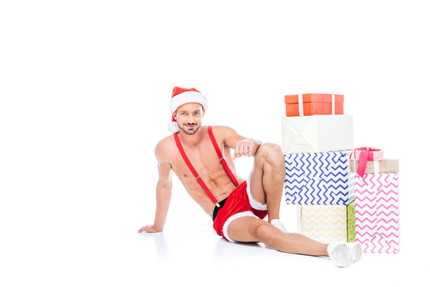 Beyaz arka plan üzerinde izole hediye kutuları yığını yakın oturan Noel şapkalı kaslı gömleksiz adam - Fotoğraf, Görsel