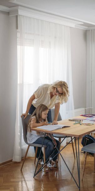 Cute blonde schoolgirl doing homework with her mother helping. - Фото, изображение