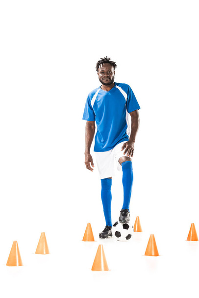 jonge Afro-Amerikaanse sportman permanent met voetbal tussen markeerdraad kegels en lachend op camera geïsoleerd op wit - Foto, afbeelding
