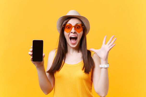 Portrét vzrušený ohromen ženy v letní klobouk, oranžové brýle držet ústa otevřít, se divíš, držet mobilní telefon s prázdnou prázdnou obrazovkou izolované na žlutém pozadí. Koncept emoce lidí - Fotografie, Obrázek