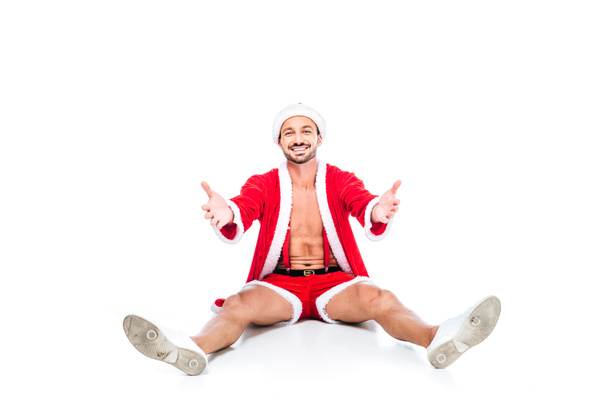 Happy svalnatý muž v kostýmu santa Clause sedí na podlaze s širokou náručí izolovaných na bílém pozadí - Fotografie, Obrázek