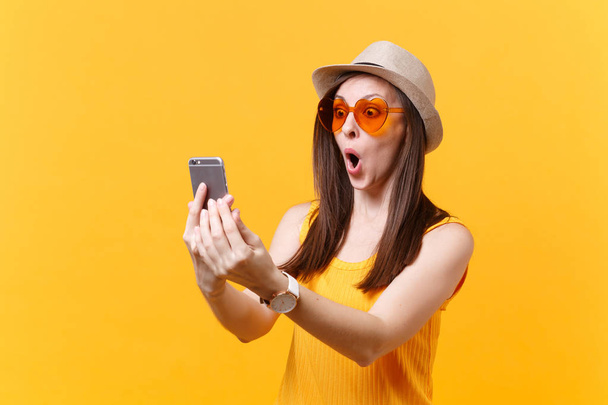 Portrait of excited amazed young woman in straw summer hat, orange glasses keeping mouth wide open, looking surprised, hold mobile phone isolated on yellow background. People sincere emotions concept - Photo, Image