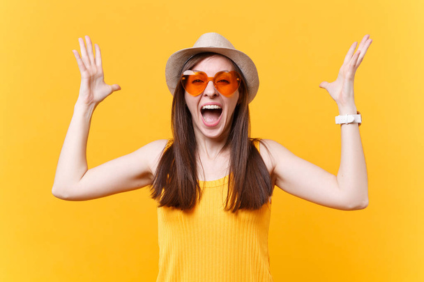 Portrait of excited screaming young woman in straw summer hat, orange glasses spreading hands, copy space isolated on yellow background. People sincere emotions, lifestyle concept. Advertising area - Photo, Image