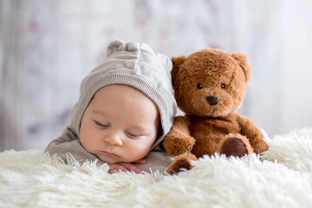 Sweet baby boy in bear overall, sleeping in bed with teddy bear stuffed toys, winter landscape behind him - Фото, изображение