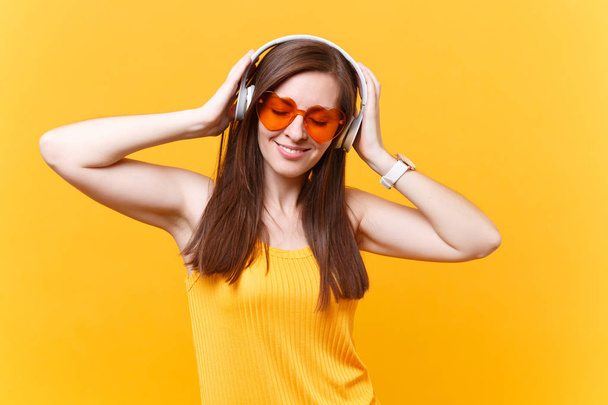 Portrait of calm smiling young woman in orange glasses listening music in headphones copy space isolated on yellow background. People sincere emotions, lifestyle concept. Advertising area. Rest relax - Foto, Bild