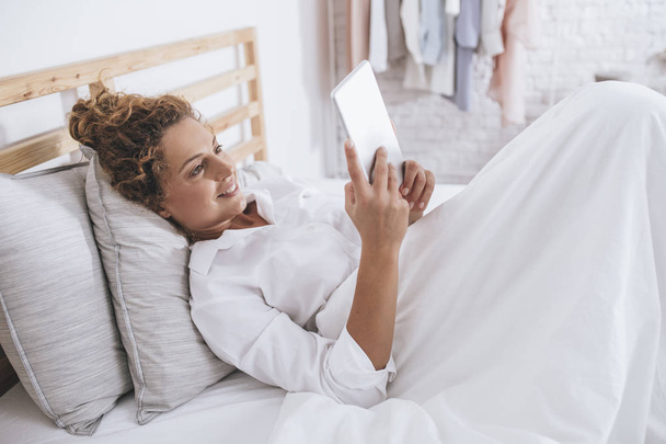 Dość uśmiechający się kaukaski kobieta leżąc w łóżku i czytania na komputerze typu tablet. - Zdjęcie, obraz