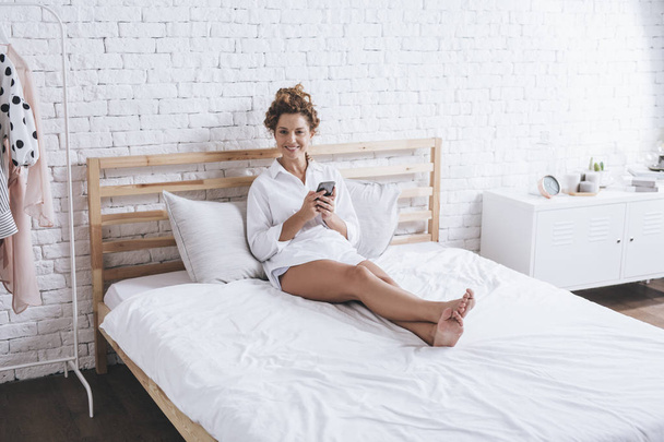 Beautiful Caucasian woman sitting on her king-size bed and typing on cell phone. - Fotoğraf, Görsel