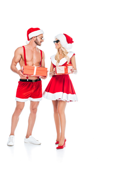 young couple in christmas hats and sunglasses holding gift boxes and looking at each other isolated on white background  - 写真・画像