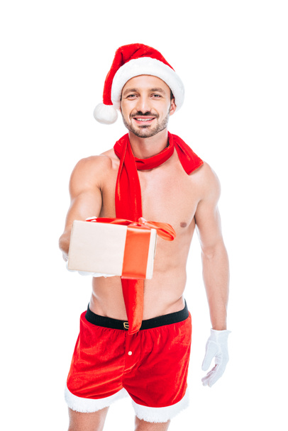 smiling shirtless muscular man in christmas hat and red scarf giving present isolated on white background - Photo, Image