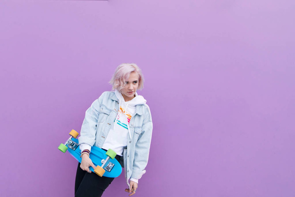 mooi meisje stijl poseren met een rots op de achtergrond van een paarse muur. Portret van een meisje-blonde die stond op een paarse achtergrond met een skate in haar armen. Copyspace - Foto, afbeelding