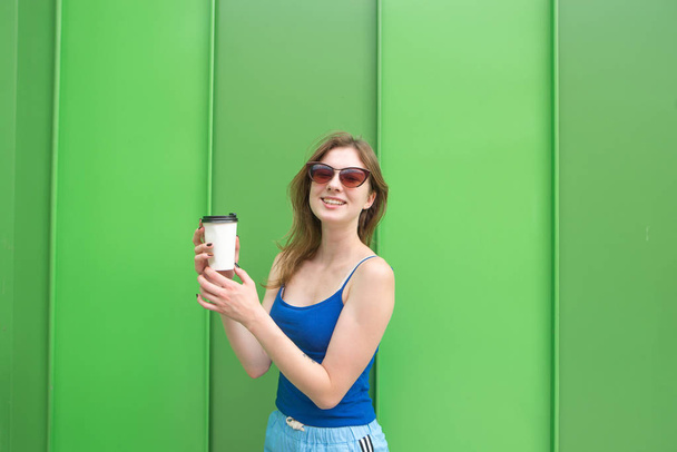 Щаслива жінка в сонцезахисних окулярах і синя футболка, що стоїть на зеленому фоні з чашкою кави в руках стіни дивиться на камеру і посміхається
. - Фото, зображення