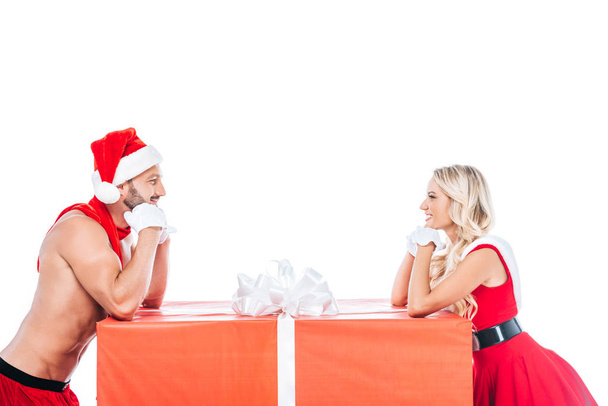 side view of dreamy couple in christmas clothes looking at each other near big gift box isolated on white background  - Zdjęcie, obraz