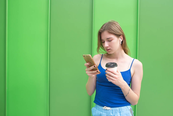 Retrato de una mujer de pie con un vaso de café en las manos, usando un smartphone y escuchando música en un auricular inalámbrico sobre un fondo de una pared verde
. - Foto, imagen