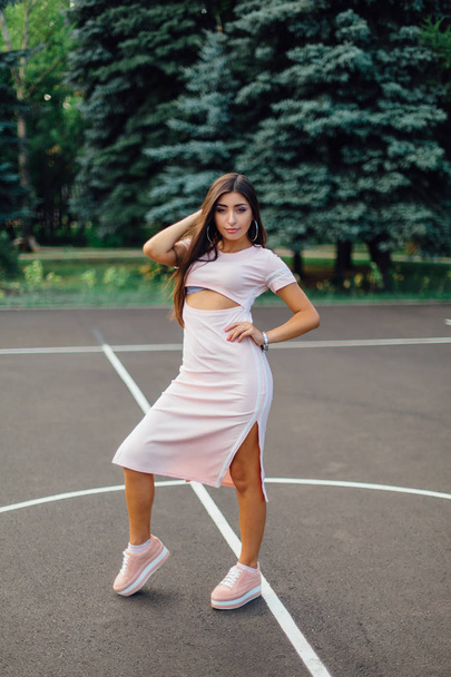 Encantadora morena vestida con un vestido rosa posando en la cancha de baloncesto al atardecer
. - Foto, Imagen