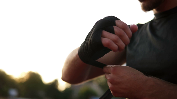 Manos de jóvenes envolviendo las manos con vendas para el entrenamiento en el día de verano al aire libre. Musculoso joven boxeador en camiseta negra. fondo arrugado
 - Metraje, vídeo