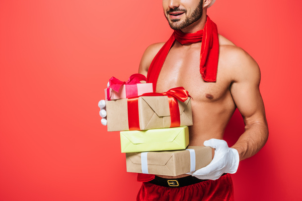 partial view of muscular shirtless sexy man in red scarf holding pile of christmas presents isolated on red background  - Foto, afbeelding