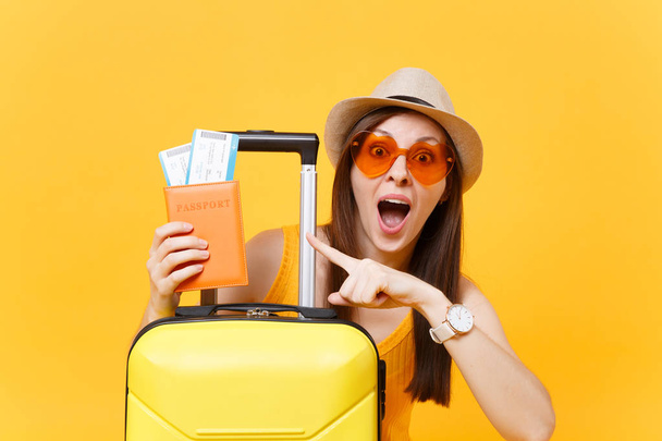 Divertida turista en ropa casual de verano, sombrero, gafas de sol naranjas con maleta, pasaporte aislado sobre fondo amarillo. Pasajero que viaja al extranjero para viajar los fines de semana de escapada. Concepto de vuelo aéreo
 - Foto, imagen