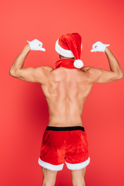 Rückansicht eines muskulösen, hemdlosen Mannes mit Weihnachtsmütze und kurzen Hosen, der Muskeln isoliert auf rotem Hintergrund zeigt  - Foto, Bild