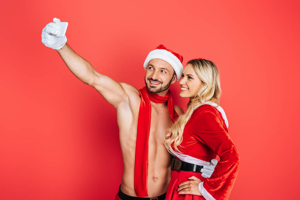 handsome muscular shirtless man in christmas hat taking selfie with girlfriend on smartphone isolated on red background - Photo, Image