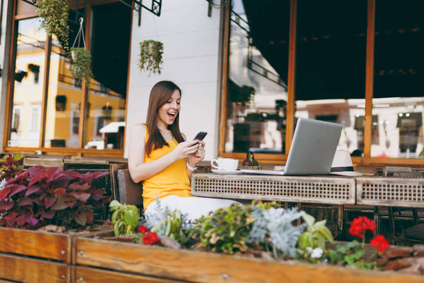 Mutlu gülümseyen kız açık havada sokak kafe Cafe ile dizüstü pc bilgisayar manifatura haber serbest zaman sırasında restoranda cep telefonu arkadaşına masada oturuyor. Mobil ofis serbest kavramı - Fotoğraf, Görsel
