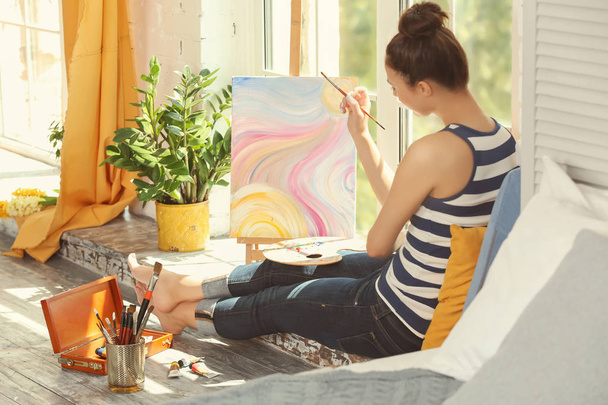 Female artist painting picture while sitting on windowsill in workshop - Foto, Imagen