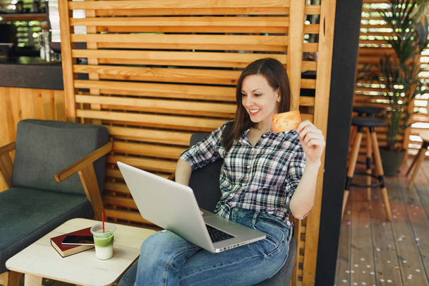 Mujer en la calle al aire libre cafetería de verano café de madera sentado trabajando en el ordenador portátil PC, mantenga la tarjeta de crédito bancaria, relajarse durante el tiempo libre. Oficina Móvil. Concepto de negocio independiente de estilo de vida
 - Foto, imagen