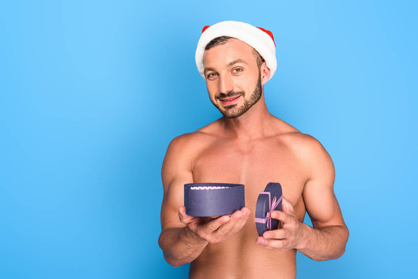 portrét shirtless svalnatého muže v klobouku vánoční drží krabičky izolované na modrém pozadí - Fotografie, Obrázek