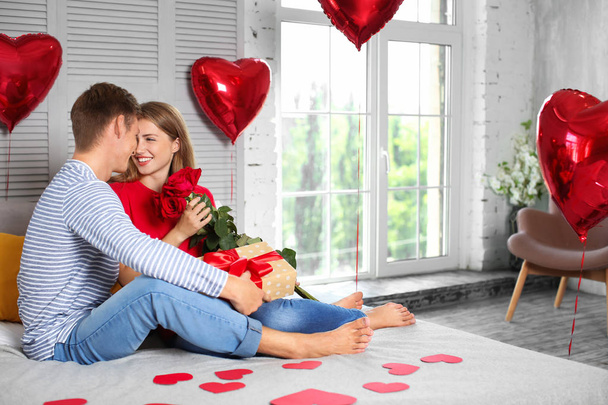 Felice giovane coppia con rose rosse e scatola regalo seduta sul letto a casa
 - Foto, immagini