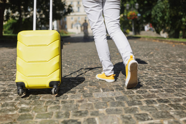 Περικομμένη εικόνα του ταξιδιώτη τουριστικά γυναίκα πόδια σε κίτρινο καλοκαίρι casual ρούχα με βαλίτσα το περπάτημα στην πόλη εξωτερική. Κορίτσι ταξίδια στο εξωτερικό για να ταξιδέψετε σε φυγή τα Σαββατοκύριακα. Τουρισμού ταξίδι ζωής - Φωτογραφία, εικόνα